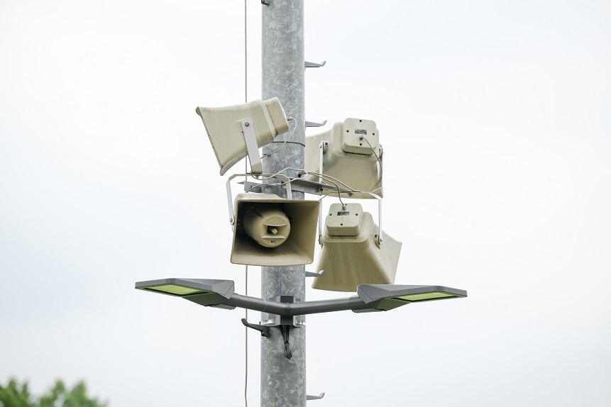 Auf dem Bild ist ein metallischer, senkrechter Mast im Außenbereich zu sehen, an dem mehrere beigefarbene Lautsprecher befestigt sind. Die Lautsprecher sind trichterförmig und in unterschiedliche Richtungen ausgerichtet, sodass sie Schall in verschiedene Richtungen abgeben können; Kabel verbinden die einzelnen Geräte miteinander. Unterhalb der Lautsprecher ist eine Straßenlaterne mit zwei flachen, rechteckigen Leuchten angebracht, die nach links und rechts zeigen. Der Hintergrund ist hell und weitgehend einheitlich, vermutlich ein bewölkter Himmel, am unteren Bildrand sind unscharf grüne Baumkronen erkennbar.
