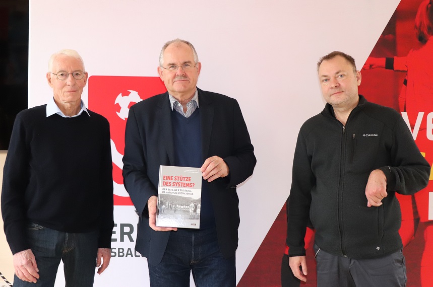 Das Foto zeigt drei Personen, die vor einer großen weißen und roten Wand mit einem grafischen Motiv stehen. In der Mitte hält eine Person ein Buch mit dem Titel „EINE STÜTZE DES SYSTEMS?". Das Buchcover ist überwiegend grau mit roten und schwarzen Schriftzügen und zeigt ein historisches Schwarz-Weiß-Foto von Fußballspielern. Die Person in der Mitte trägt ein dunkles Sakko über einem blauen Pullover und einem Hemd. Die Person links ist in einem dunklen Pullover mit weißem Hemd gekleidet, während die Person rechts eine dunkle Outdoorjacke trägt. Im Hintergrund ist ein Teil eines Logos sowie rote grafische Elemente sichtbar, die auf einen sportlichen Kontext hinweisen.