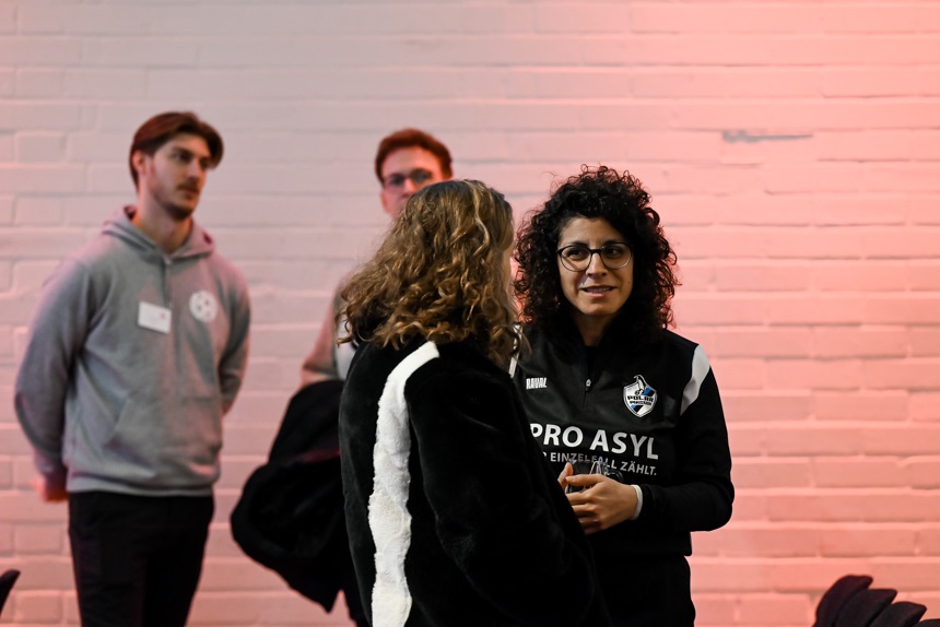 In einem hellen Raum mit einer weiß gestrichenen Backsteinwand stehen mehrere Personen zusammen. Im Vordergrund unterhalten sich zwei Menschen miteinander. Eine Person trägt eine schwarze Jacke mit einem weißen Streifen auf dem Rücken, die andere eine schwarze Sportjacke mit dem Aufdruck „PRO ASYL – Ein Recht, das zählt.“ Im Hintergrund stehen zwei weitere Personen etwas abseits und beobachten die Szene. Der Raum ist warm beleuchtet, mit einem rötlichen Lichtschein auf der Wand. Die Atmosphäre wirkt ruhig und konzentriert, als befänden sich die Personen in einer Gesprächssituation oder bei einer Veranstaltung.
