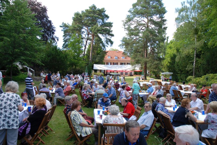 Menschen sitzen im Garten an Tischen, essen und unterhalten sich bei einem großen Fest vor einem Gebäude im Grünen.