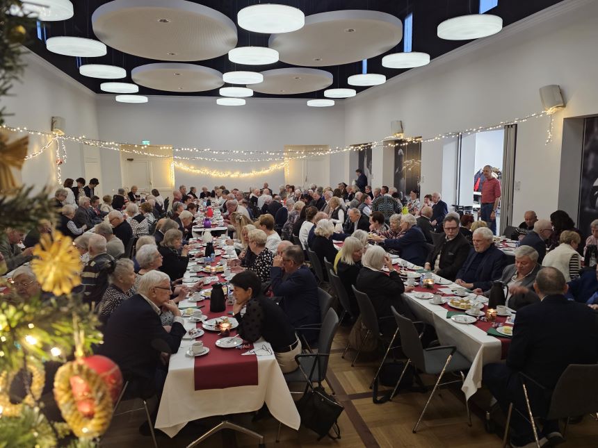 Festlich geschmückter Saal, in dem viele Menschen an langen, gedeckten Tischen gemeinsam Kaffee und Kuchen genießen.