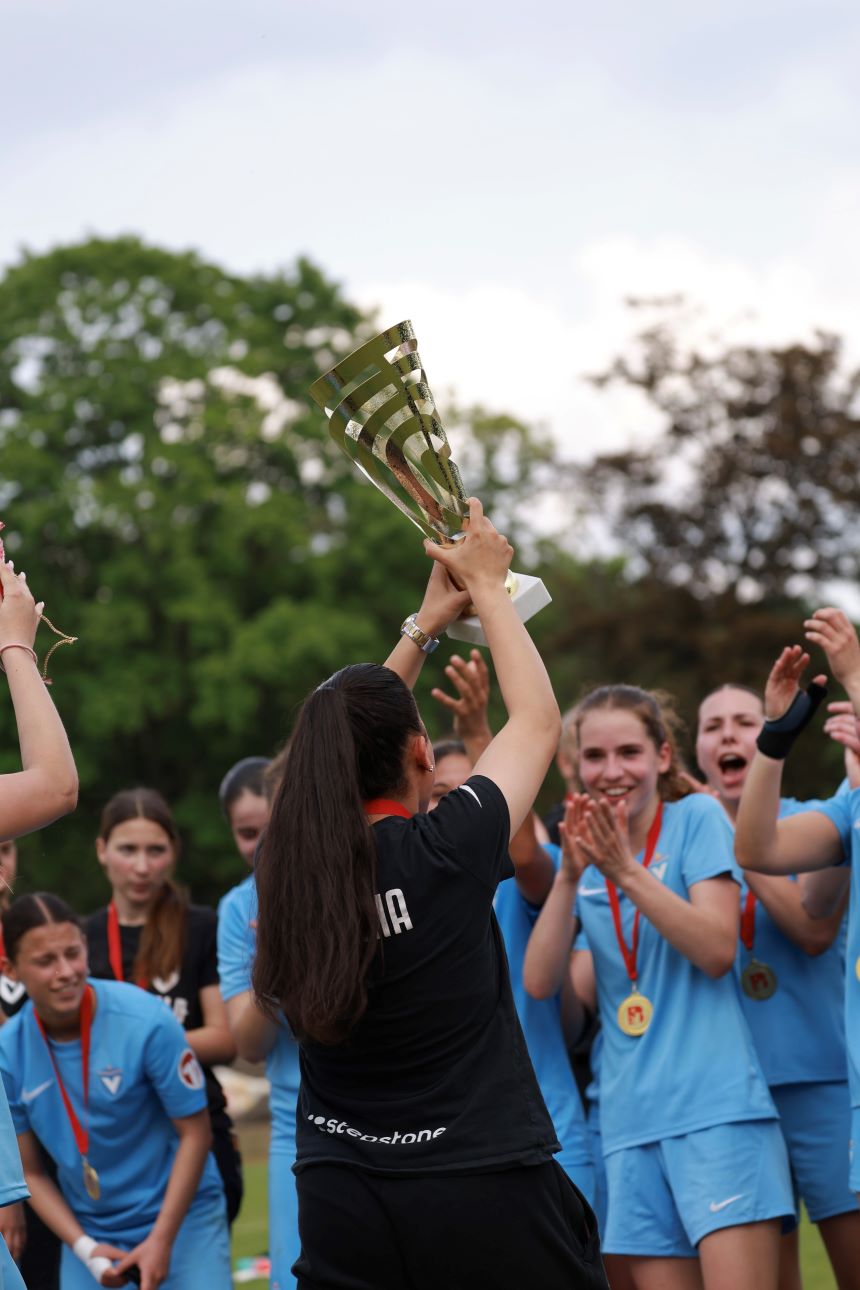 Person hebt Pokal vor Team mit Medaillen; helle Trikots, Bäume im Hintergrund, sonniger Himmel auf dem Fußballfeld.