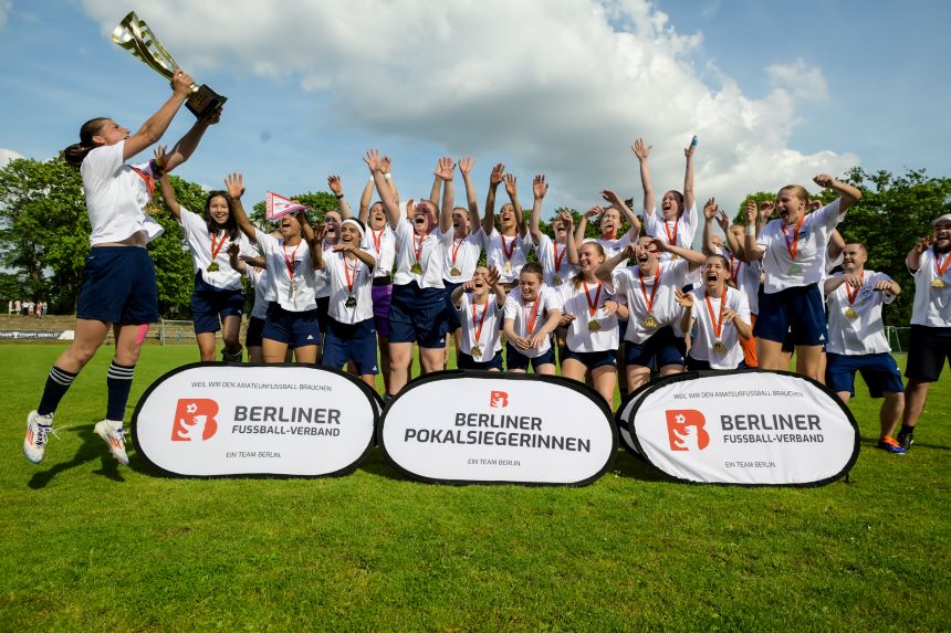 Gruppe feiert auf Fußballplatz, hebt Pokal, trägt Medaillen; Banner „Berliner Pokalsiegerinnen“, blauer Himmel im Hintergrund.