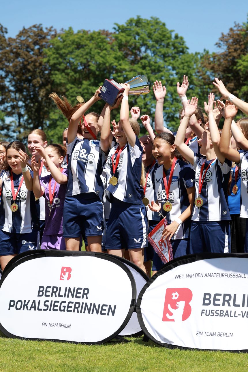 Jubelnde Fußballmannschaft in blau-weißen Trikots hebt Pokal; viele tragen Medaillen, Banner „Berliner Pokalsiegerinnen“ im Vordergrund.