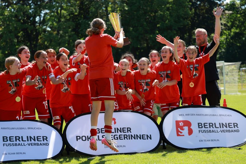 Gruppe junger Fußballspielender in roten Trikots jubelt auf Spielfeld; Person hält Pokal hoch, Banner „Berliner Fußball-Verband“ im Vordergrund.