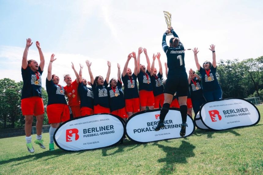 Team auf Fußballfeld hebt gemeinsam einen Pokal; alle tragen Medaillen, Banner „Berliner Pokalsiegerinnen“, sonniger Himmel im Hintergrund.