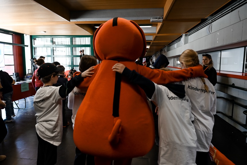 Auf dem Foto steht eine große orangefarbene Maskottchenfigur mit dem Rücken zur Kamera und hat beide Arme weit ausgestreckt, sodass sie mehrere Kinder gleichzeitig umarmt. Die Kinder tragen weiße T‑Shirts mit der Aufschrift „Kinderreporter“ und stehen eng beisammen. Die Szene spielt in einer hellen Halle mit großen Fenstern, in der weitere Menschen und Stände im Hintergrund zu sehen sind. Die Atmosphäre wirkt lebendig und fröhlich, als würde eine gemeinsame Aktion oder Veranstaltung stattfinden.