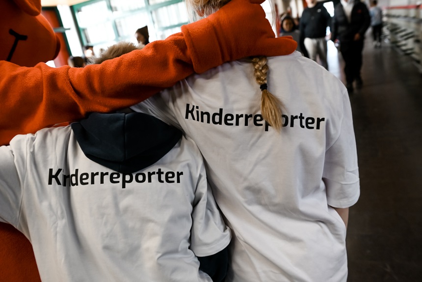 Auf dem Foto stehen zwei Kinder nebeneinander, beide tragen weiße T‑Shirts mit der Aufschrift „Kinderreporter“ auf dem Rücken. Eine große Figur mit orangefarbenem Kostüm legt einem der Kinder einen Arm freundschaftlich über die Schulter. Die Szene spielt in einem hellen Innenraum, in dem weitere Personen im Hintergrund zu sehen sind. Die Atmosphäre wirkt lebhaft und fröhlich.
