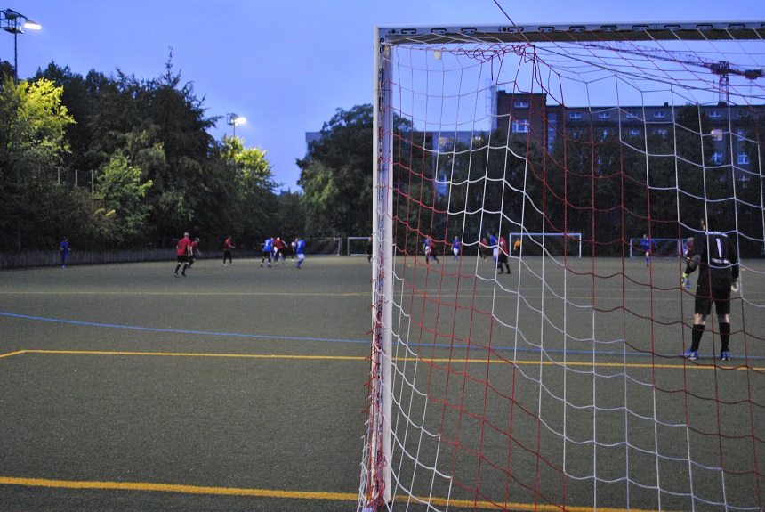 The photo shows a soccer pitch as dusk falls. In the foreground is a goal with a net made of red and white strings. A goalkeeper in dark clothing stands in the goal area on the right. In the background, two teams are playing against each other: some players are wearing red shirts, others blue. The game takes place on a large artificial turf pitch surrounded by trees. Behind the trees, multi-storey residential buildings and a construction crane can be seen. Several floodlight masts illuminate the pitch and provide bright, even lighting for the scene.