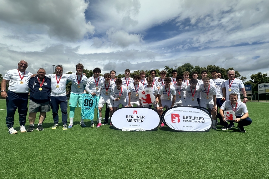 A youth soccer team in white jerseys, holding medals and the championship trophy, on the soccer field behind a sign for the Berlin Championship.
