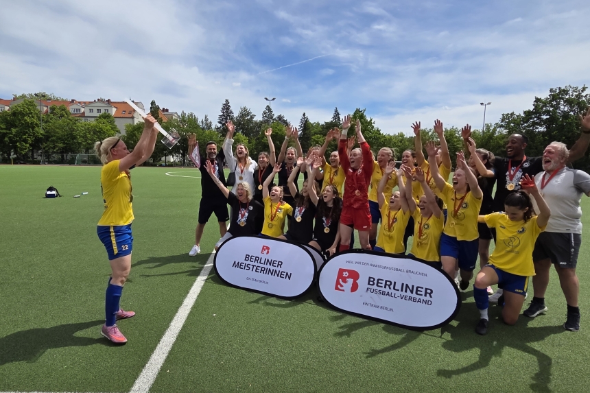 A sports team in yellow jerseys with medals on the soccer field; one person is holding up a trophy, while others are celebrating in front of signs for the Berlin Championship.