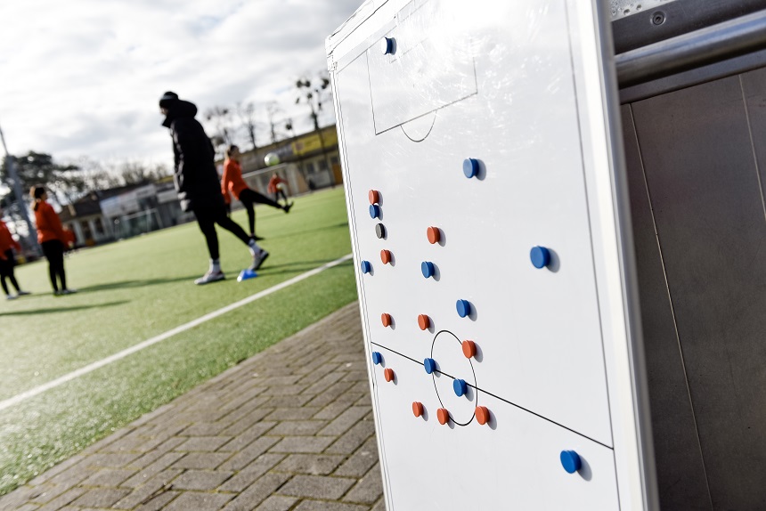 Auf dem Bild steht im Vordergrund eine weiße Taktiktafel mit vielen kleinen Magnetpunkten in Rot und Blau, die eine Fußballaufstellung zeigen. Im Hintergrund sieht man ein paar Personen auf einem Fußballplatz. Sie tragen Sportkleidung und bewegen sich über das Feld. Der Fokus des Bildes liegt auf der Tafel, nicht auf den Personen.
