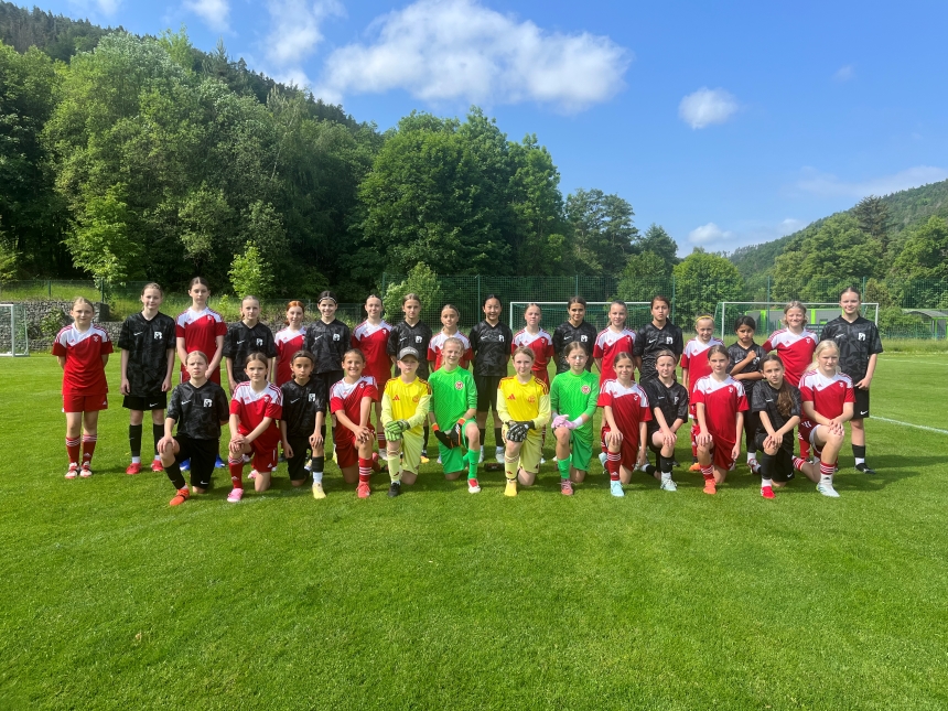 Gruppe junger Fußballspielender in roten und schwarzen Trikots auf Rasen, in Reihen aufgestellt, mit Toren, Bäumen und blauem Himmel im Hintergrund.