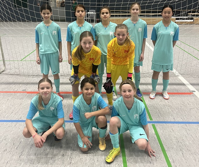 A youth soccer team poses in front of an indoor gate in a sports hall. The children are wearing predominantly turquoise jerseys, shorts and socks. Two players in the middle row are wearing striking yellow goalkeeper clothing, one of them in yellow with an orange pattern, the other in bright yellow kit. The front row consists of three children posing in a squatting position, while the remaining team members stand behind them. 