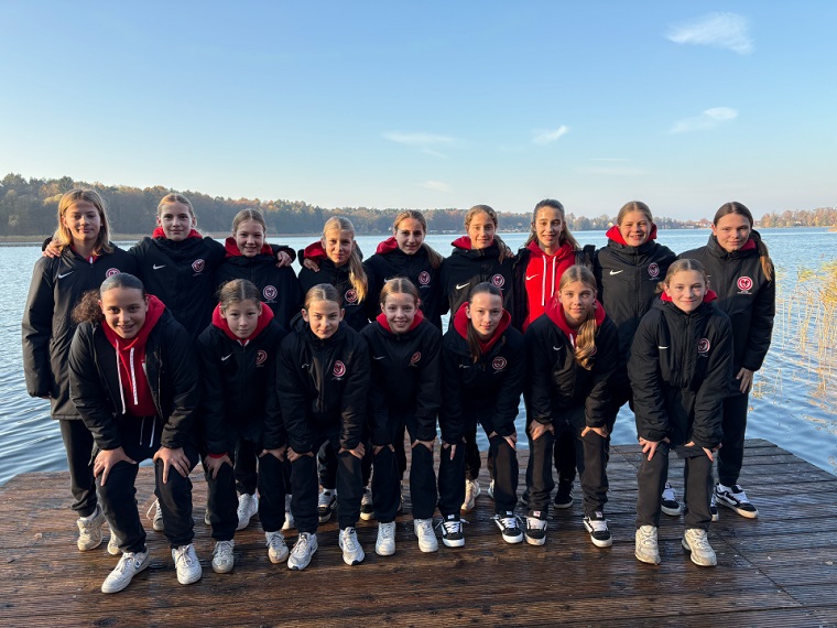 Das Foto zeigt eine Gruppe von 16 Personen, die auf einem hölzernen Steg am Ufer eines Sees stehen. Die Gruppe ist in zwei Reihen angeordnet: eine vordere Reihe in leicht gebückter Haltung und eine hintere Reihe aufrecht stehend. Alle tragen einheitliche schwarze Sportjacken mit roten Kapuzenpullovern darunter sowie schwarze Hosen und Sportschuhe. Auf den Jacken ist ein rundes Vereinslogo in Weiß und Rot sichtbar. Im Hintergrund ist ein ruhiger See zu sehen, umgeben von herbstlich gefärbten Bäumen unter einem klaren blauen Himmel. Das Licht wirkt wie spätes Vormittags- oder Nachmittagslicht, wodurch warme Farbtöne entstehen.