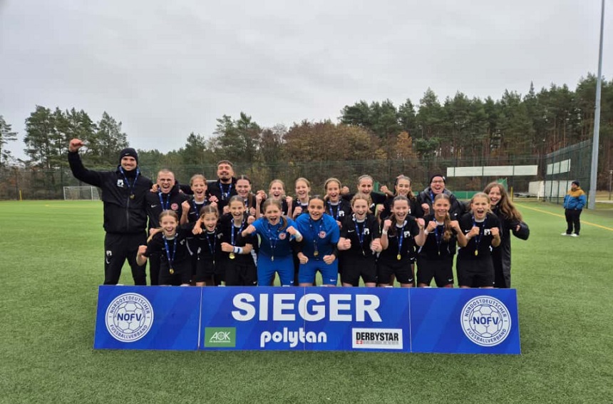 Das Foto zeigt eine Fußballmannschaft auf einem Kunstrasenplatz. Die Gruppe steht dicht beieinander hinter einem großen blauen Schild mit der weißen Aufschrift „SIEGER“. Auf dem Schild sind außerdem Logos des Nordostdeutschen Fußballverbands (NOFV) sowie der Sponsoren „AOK“, „polytan“ und „DERBYSTAR“ zu sehen. Alle Personen tragen schwarze Sportkleidung, einige haben Medaillen um den Hals. In der Mitte hält eine Person einen Pokal. Im Hintergrund sind Bäume mit herbstlichem Laub, ein hoher Zaun und ein Flutlichtmast erkennbar. 