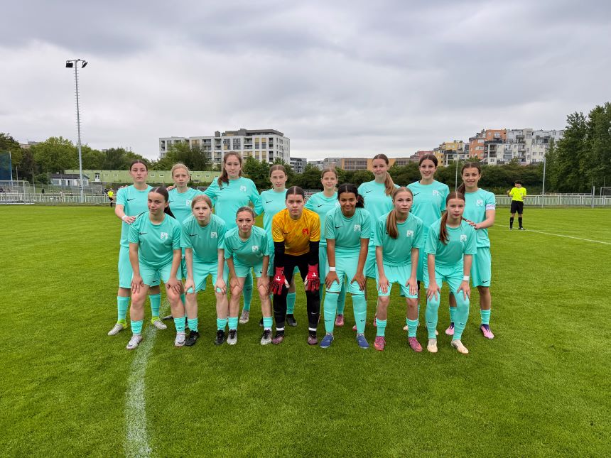 Gruppe junger Fußballspielender in hellblauen Trikots auf Rasen, vor Wohnhäusern; mittig Person in gelbem Torwartoutfit, bewölkter Himmel.