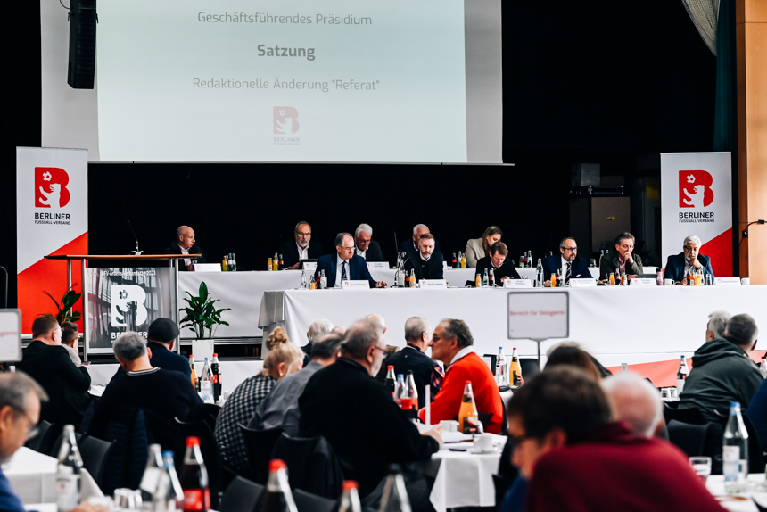 A conference with several people at the podium in front of a screen that reads 'Executive Presidium - Statutes - Editorial change "Referat"'. In the foreground, participants are sitting at tables with drinks. Banners with the Berliner logo are visible on the left and right.
