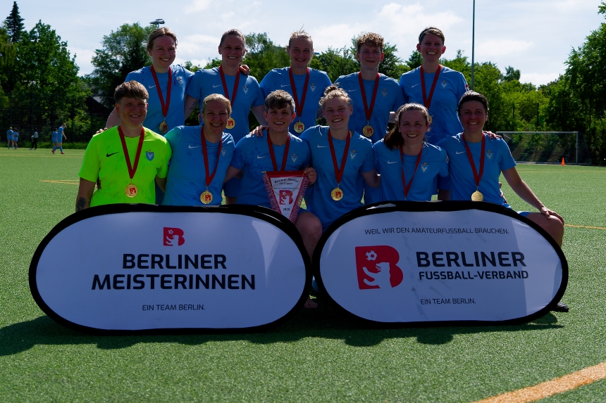 A sports team in light blue jerseys, wearing medals and carrying a championship pennant, on the sports field in front of signs for the Berlin Championship.