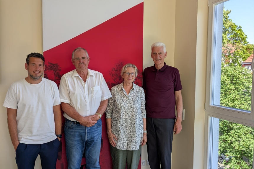 Four people are standing in a bright room in front of a red-and-white wall; on the right is a large window with a view of green trees.