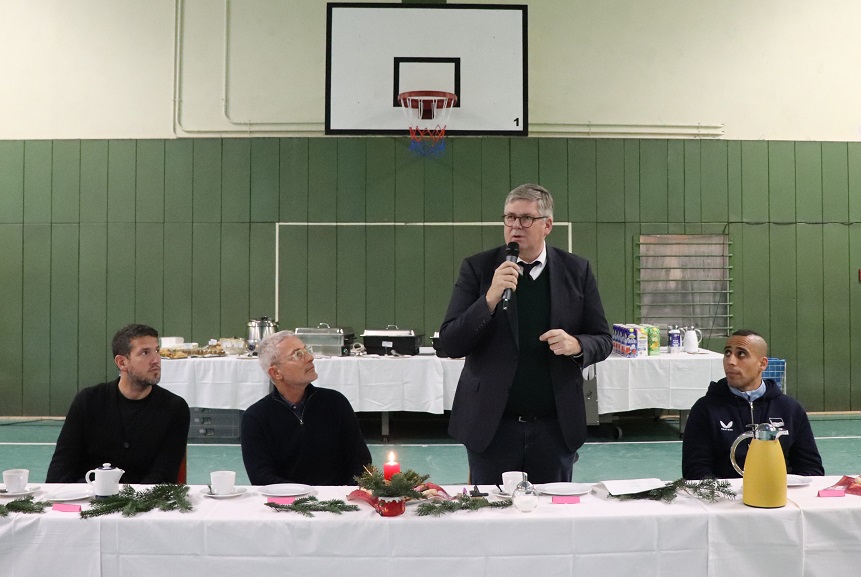 In a sports hall, a person stands behind a long table and speaks into a microphone. On the table are cups, plates, a yellow thermos flask and some Christmas decorations with a candle. In the background are more tables with drinks and food as well as a basketball hoop on the wall.