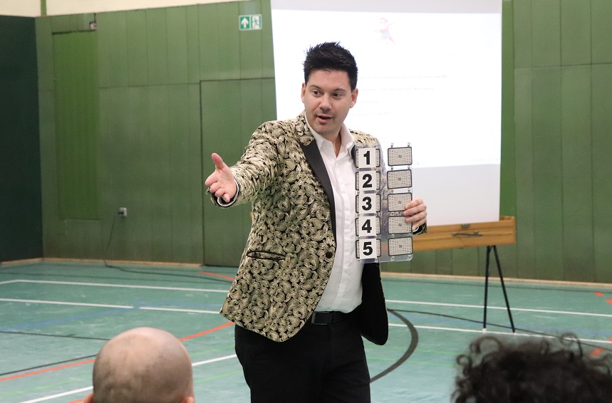 Eine Person steht in einer Sporthalle vor einer Projektionsfläche. Die Person trägt ein gemustertes Sakko und hält ein längliches Objekt mit fünf übereinander angeordneten Tafeln, nummeriert von 1 bis 5. Er ist ein Zauberer. Die linke Hand ist nach vorne ausgestreckt, als würde die Person das Publikum ansprechen. Im Vordergrund sind Köpfe von sitzenden Personen sichtbar.