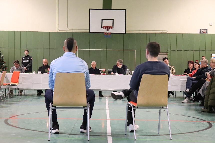 In a sports hall, two people are sitting on chairs with their backs to the camera. In front of them is a long table where several people are sitting and listening. In the background is a basketball hoop on the wall. On the right of the picture, other people are sitting in a row, on the left are some empty chairs.