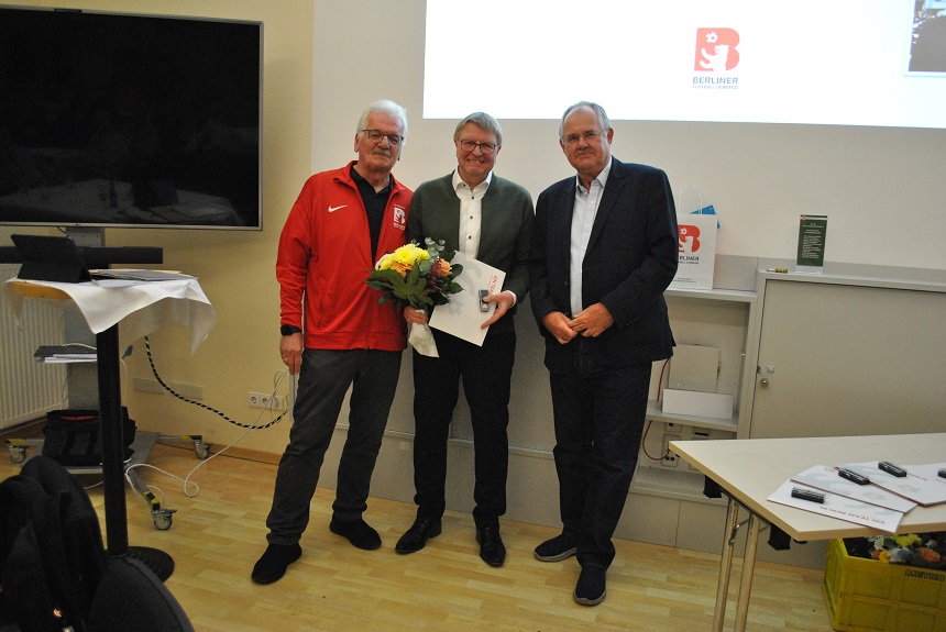 Drei erwachsene Personen stehen nebeneinander in einem hellen Seminarraum. Die Person in der Mitte hält einen Blumenstrauß und eine Urkunde in den Händen. Links steht eine Person in einer roten Sportjacke, rechts eine Person in dunkler Kleidung. Im Hintergrund sind eine Präsentationsfläche mit einem Logo sowie ein großer Bildschirm zu sehen. Neben den Personen stehen Tische mit Unterlagen und Technik.