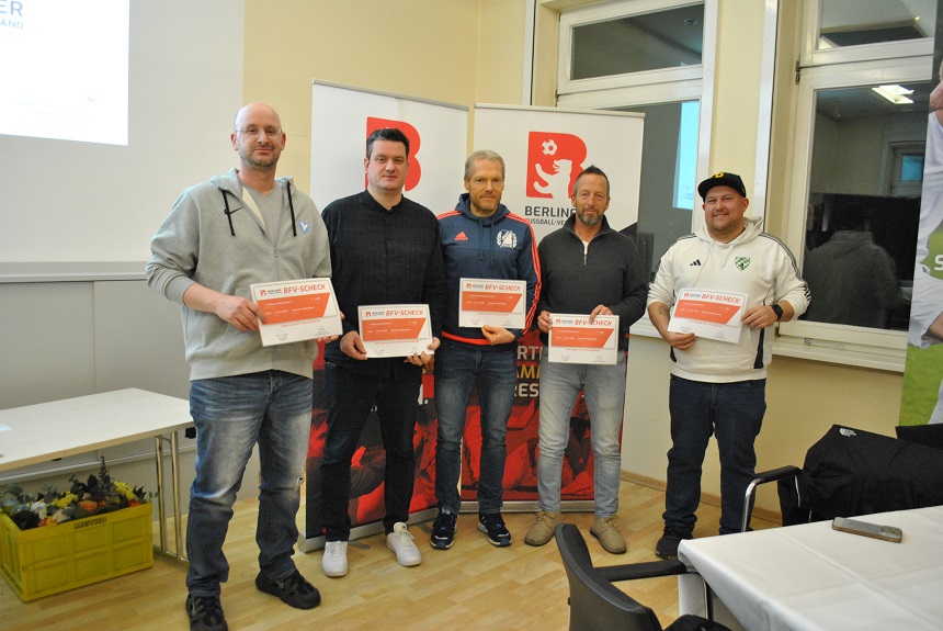 Fünf erwachsene Personen stehen nebeneinander in einem Veranstaltungsraum und halten jeweils eine Urkunde in den Händen. Hinter ihnen sind Roll‑ups mit dem Logo des Berliner Fußball‑Verbands zu sehen. Die Personen sind unterschiedlich gekleidet, unter anderem in sportlicher Freizeit‑ und Vereinskleidung. Der Raum ist hell, mit Fenstern, einem Tisch im Vordergrund und weiterem Veranstaltungsmaterial im Hintergrund. 