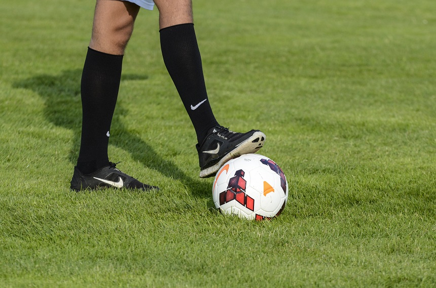 Auf dem Bild steht eine Person auf einer grünen Rasenfläche. Ein Fuß ruht auf einem Fußball, der weiß ist und rote sowie schwarze Muster hat. Die Person trägt schwarze Sportschuhe, schwarze Stutzen und eine kurze helle Hose. Foto: Sandra Ritschel
