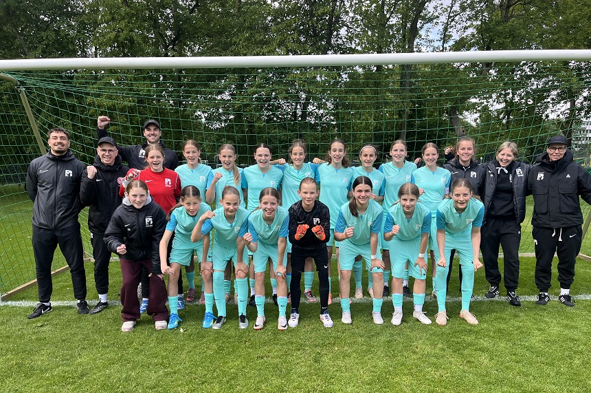 Die Berliner U14-Auswahl posiert geschlossen vor einem Fußballtor auf einem Rasenplatz. Das Team trägt einheitlich helle, türkisfarbene Trikots und steht in zwei Reihen – eine kniende Reihe vorne und eine stehende dahinter. In der Mitte ist eine Torhüterin in dunkler Ausrüstung mit Handschuhen platziert. Seitlich neben der Mannschaft stehen mehrere Mitglieder des Trainer- und Betreuerteams in dunkler Sportkleidung. Einige Teammitglieder zeigen eine geballte Faust in Richtung Kamera. Im Hintergrund sind Bäume und ein dichter grüner Zaun zu sehen, die dem Bild einen natürlichen Rahmen geben.