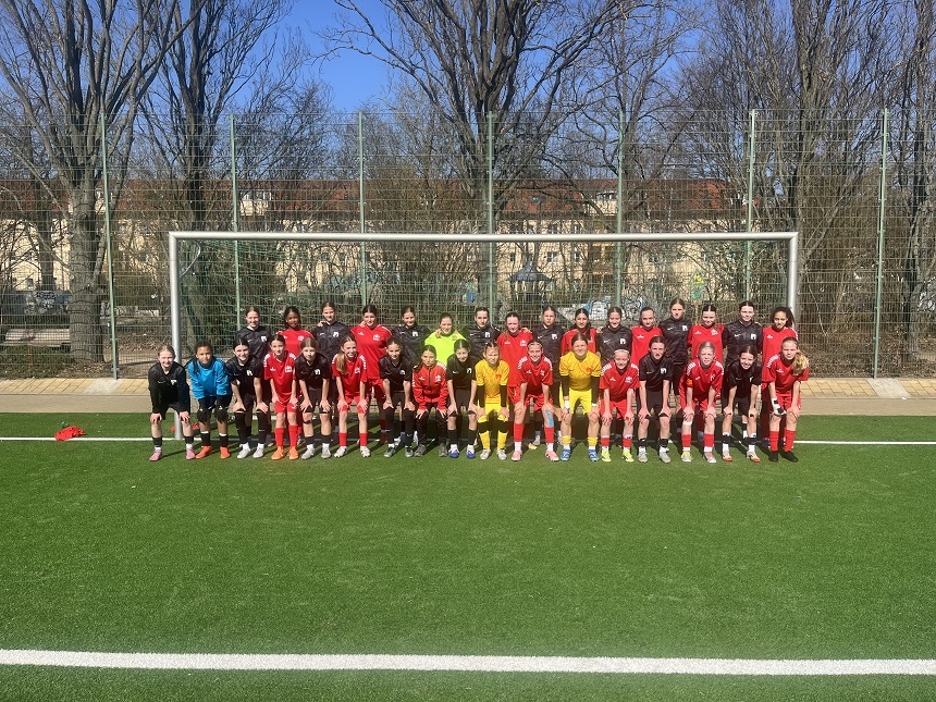 Auf einem sonnigen Fußballplatz steht eine große Gruppe von Spielenden in mehreren Reihen direkt vor einem Tor. Die Gruppe trägt unterschiedliche Trikotsätze: Ein Teil ist in roten Trikots mit roten Hosen gekleidet, ein anderer Bereich in dunklen Trikots, und einige Personen tragen gelbe oder hellblaue Torwartkleidung. Alle stehen eng nebeneinander auf dem grünen Kunstrasen, wobei die vordere Reihe leicht in die Knie geht, während die hintere Reihe aufrecht steht. Im Hintergrund befindet sich ein Metallzaun, dahinter Bäume ohne Laub sowie Gebäude, die nur teilweise durch die Äste sichtbar sind. Der Himmel ist klar und blau, und die Sonne wirft deutliche Schatten nach hinten. Die Szene wirkt wie ein gemeinsames Gruppenfoto zweier oder mehrerer Teams nach einem Spiel oder Trainingsereignis.