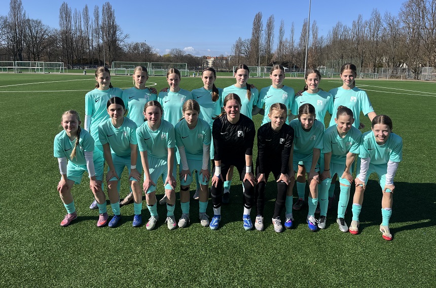 Eine Fußballmannschaft steht auf einem grünen Kunstrasenplatz, umgeben von kahlen Bäumen unter einem klaren blauen Himmel. Die Gruppe trägt einheitliche türkisfarbene Trikots, kurze Hosen und Stutzen; zwei Personen in der vorderen Reihe tragen dunklere Trainingskleidung. Die vordere Reihe kniet oder hockt, während die hintere Reihe steht. Im Hintergrund sind weitere Fußballfelder, weiße Linienmarkierungen und ein hoher Flutlichtmast zu sehen. Das Team wirkt ordentlich aufgestellt, als würde ein offizielles Mannschaftsfoto aufgenommen.