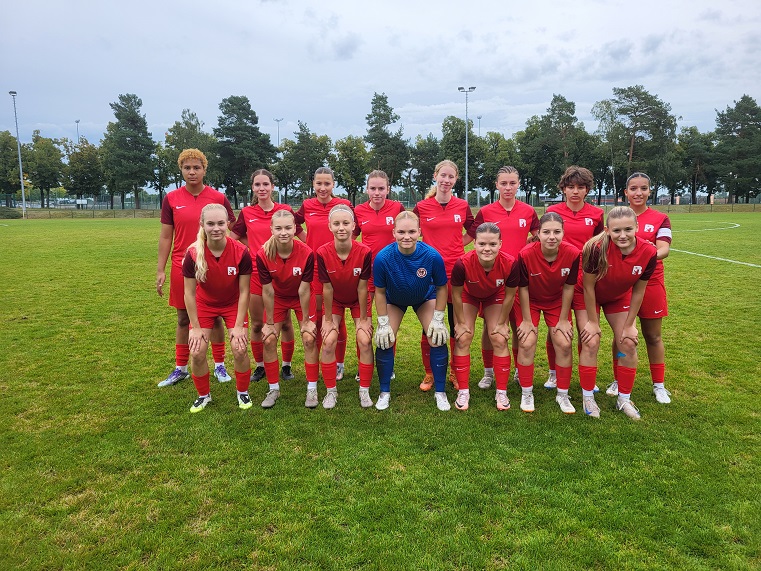 A football team is standing together on a grass pitch, all team members are wearing red shirts and soccer boots. Eight players are kneeling in the front row, one player in a blue goalkeeper jersey is sitting in the middle, while seven other team members and a coach are standing in the back row. Trees and a playing field can be seen in the background.