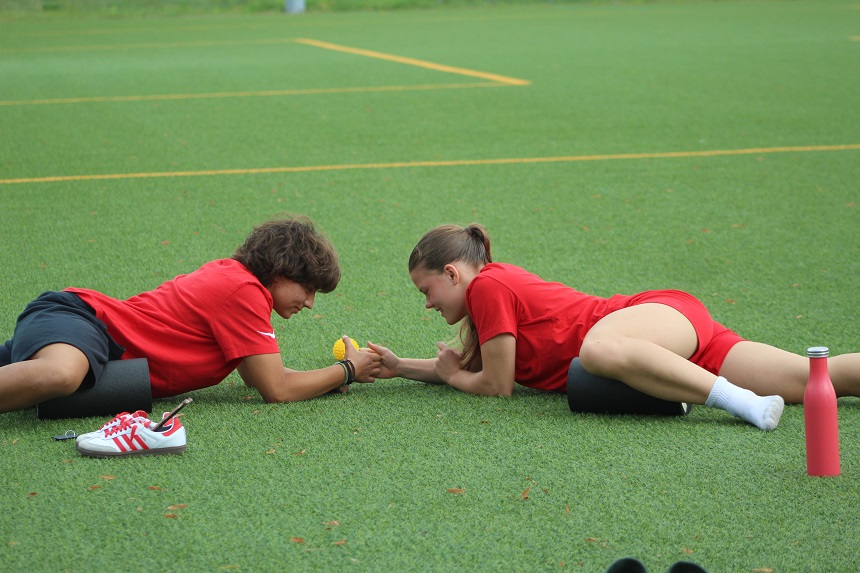 Two teenagers in red sports outfits lie opposite each other on an artificial pitch, leaning on their forearms, with a yellow ball between them. Next to them are sports shoes, a water bottle and foam rollers.