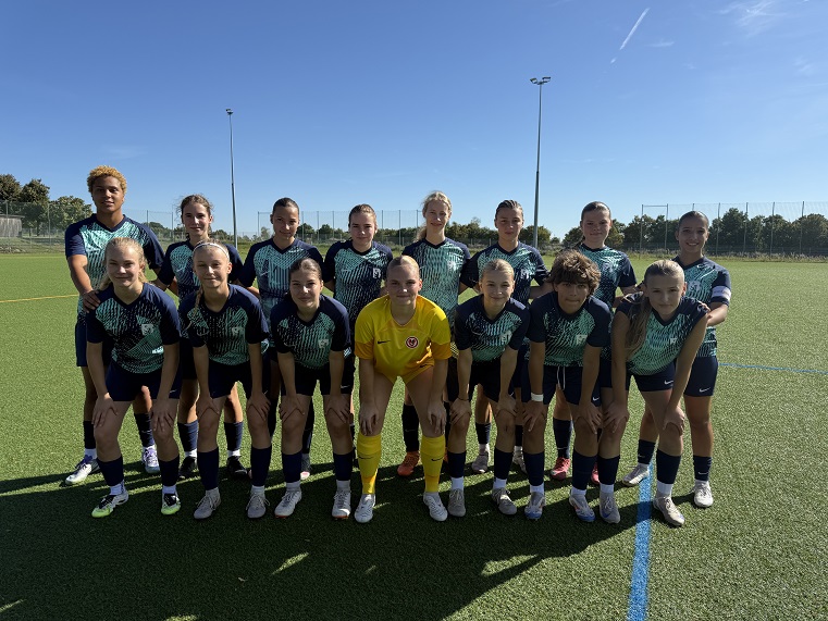 A football team stands for a group photo on an artificial turf pitch. The players are wearing dark blue jerseys with light patterns, black shorts and socks, while a player in a yellow goalkeeper's jersey stands in the front row. In the background, trees, a fence and floodlight masts can be seen under a blue sky.